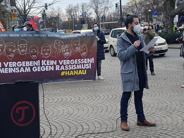 Ein Mann im grauen Dufflecoat trägt Mundschutz, während er bei einer Demonstration einen Text vorliest.