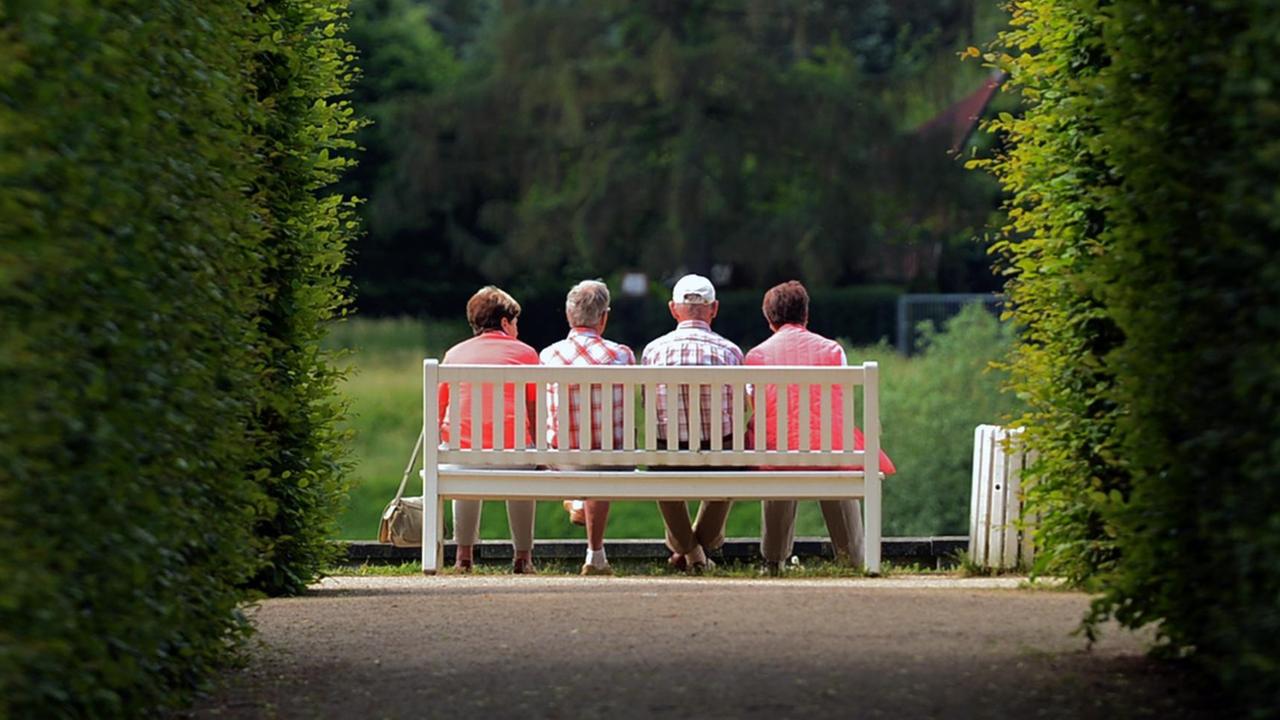 Mehrere ältere Menschen sitzen auf einer Park-Bank. 