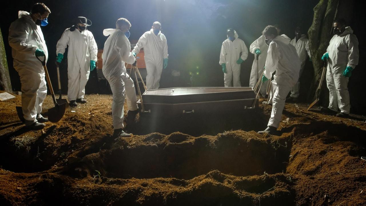 Mitarbeiter eines Friedhofs tragen einen Sarg, um eine an Covid-19 verstorbene Person zu beerdigen. Es ist Nacht, im Hintergrund sind Lichtstrahler aufgestellt, man sieht mehrere ausgehobene Gräber hintereinander.