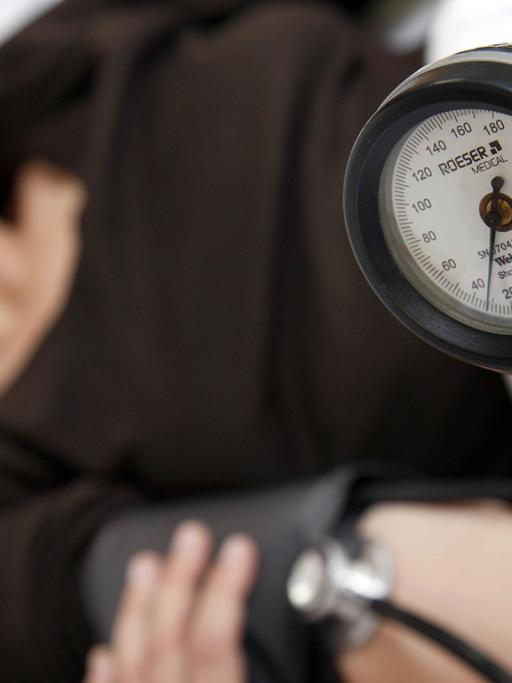 Ein Frau mit schwarzem Oberteil auf einer Liege; im Vordergrund ist die Messuhr eine Blutdruckgeräts zu sehen, das eine andere Person, die nicht im Bild ist, in der Hand hält.