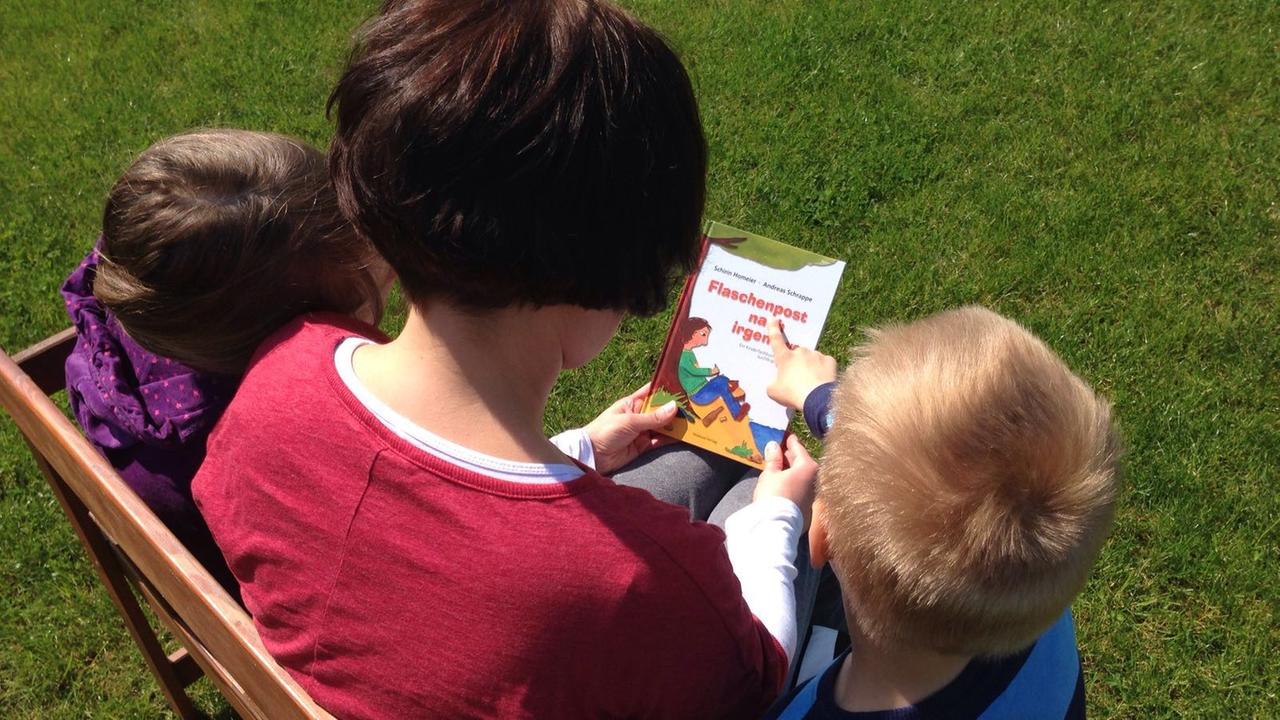 Eine Mutter sitzt mit ihren zwei Kindern auf einer Bank und hält ein Buch in den Händen.