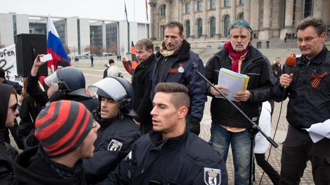 Vier Reichsbürger stehen hinter einem Mikrofoständer, einer von ihnen hält das Mikro. Drei Polizisten stehen davor, sie riegeln die Kundgebung gegen Gegendemonstranten ab.