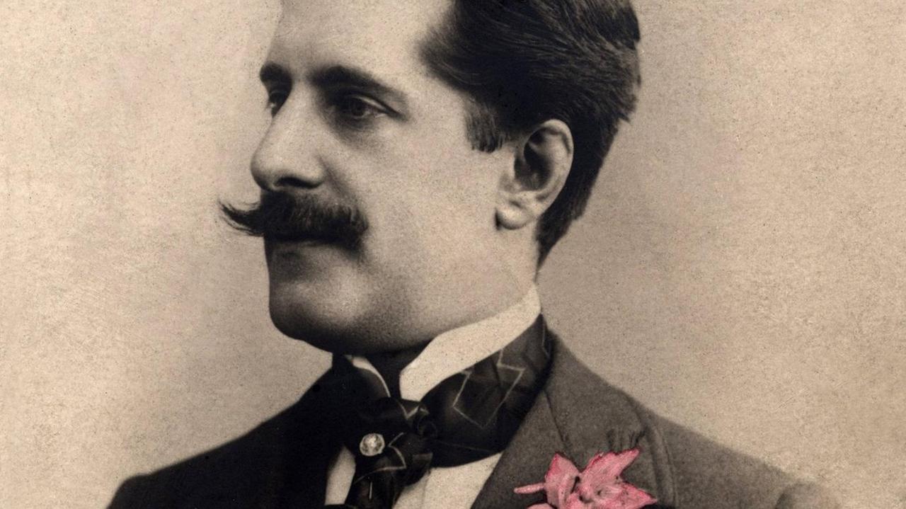 Historische Fotografie von 1895 des Komponisten Albert Roussel, der einen großen Schnauzbart und eine nachkolorierte, rosafarbene Blume am Kragen trägt.
