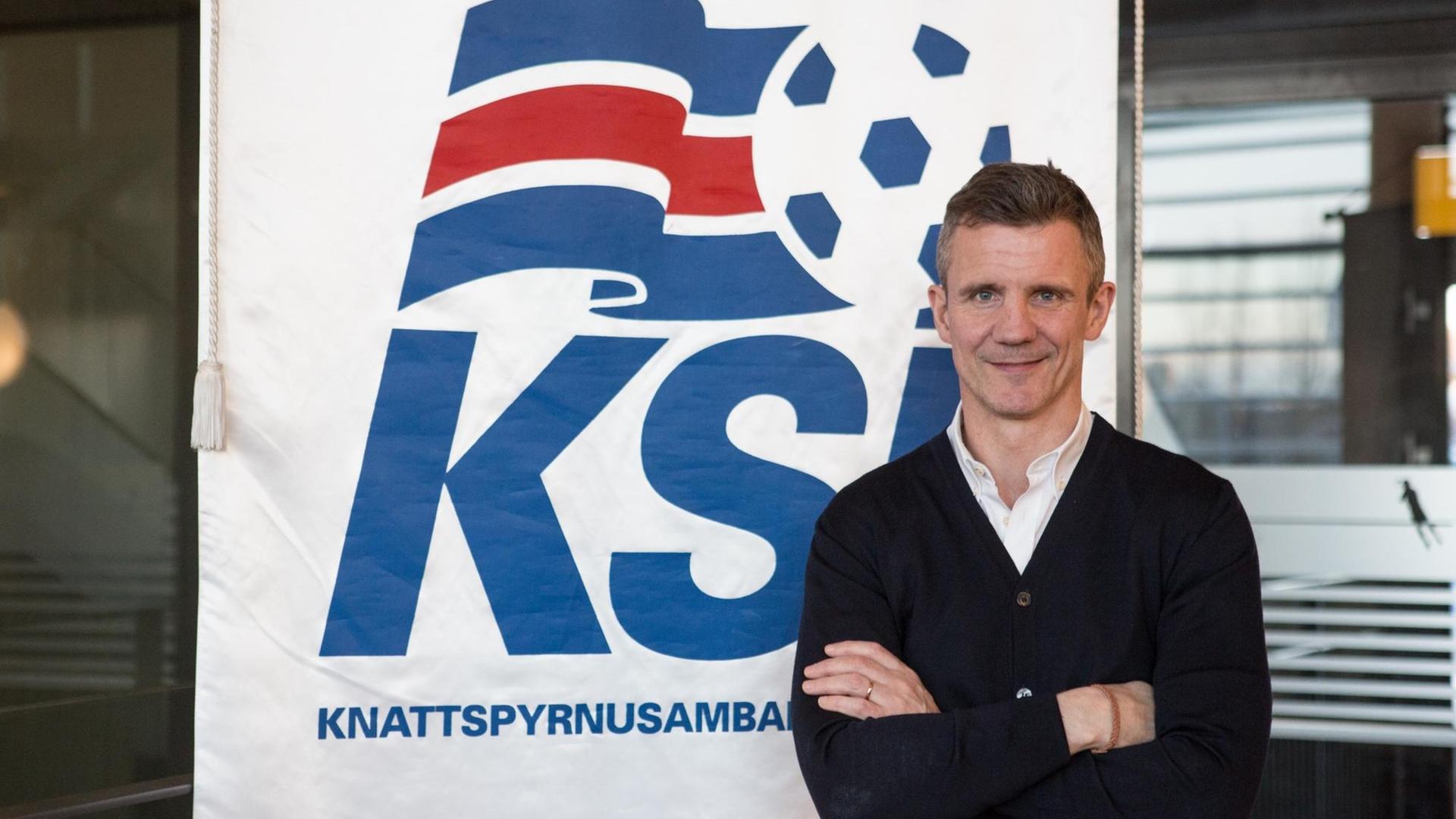 Guðni Bergsson steht mit verschränkten Armen vor einem Plakat des Verbandes KSÍ, er lächelt und trägt ein weißes Hemd und einen schwarzen Strickpulli