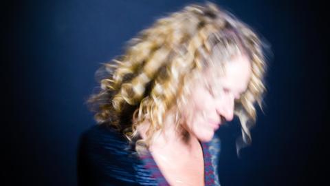 Eine Frau mit blondem Lockenkopf ist durch Bewegung unscharf vor einem dunklen Hintergrund zu sehen und schaut nach unten.