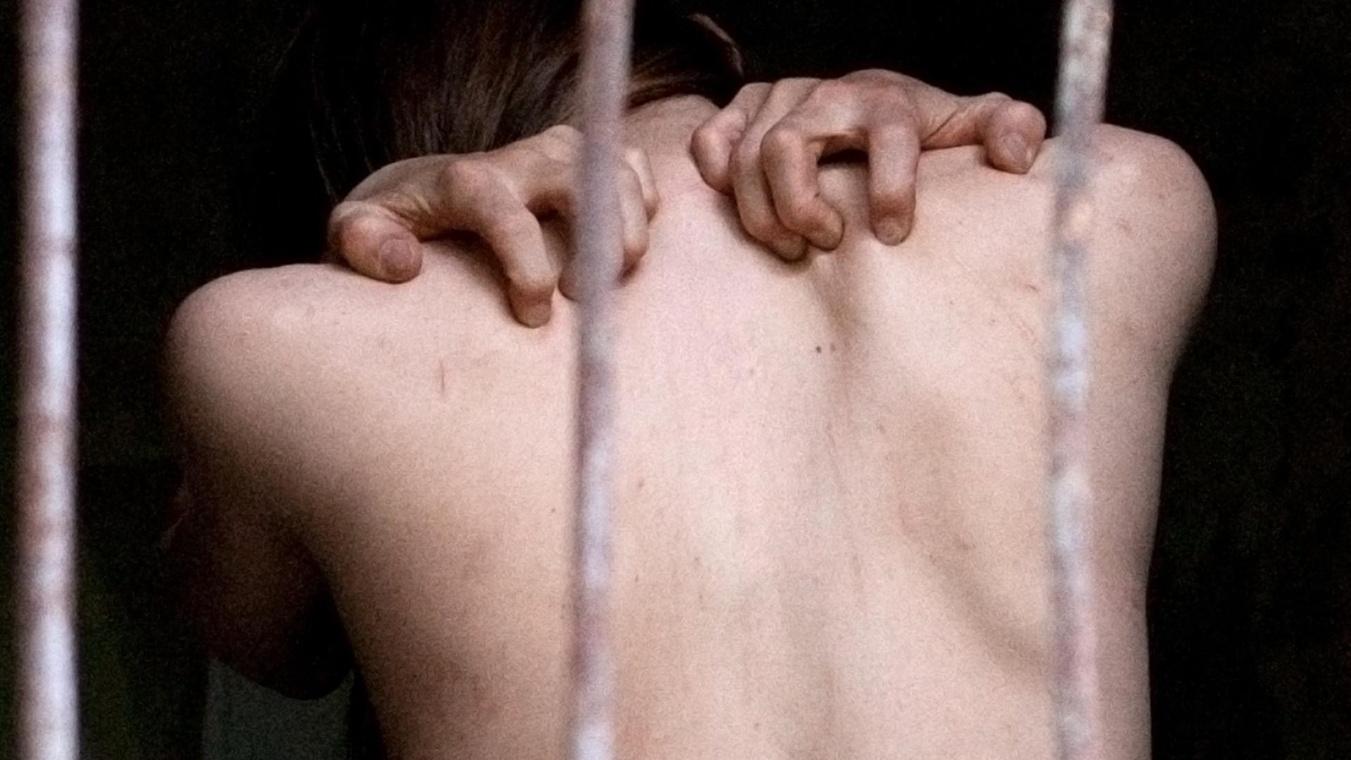 Hinter drei vertikalen Metallstangen ist ein nackter oberer Rückenausschnitt eines Menschen zu sehen, der seine Finger in die Schultern presst
