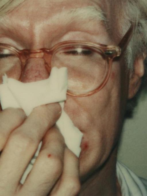 Das Bild zeigt ein Polaroid-Foto auf dem Andy Warhol sich die Nase putzt