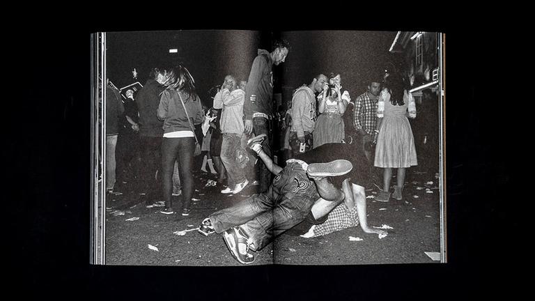 Das Bild zeigt eine Doppelseite aus einem Fotoband vor schwarzem Hintergrund: Auf der Münchner Wiesn sind liegen zwei betrunkene Männer übereinander auf dem Boden.