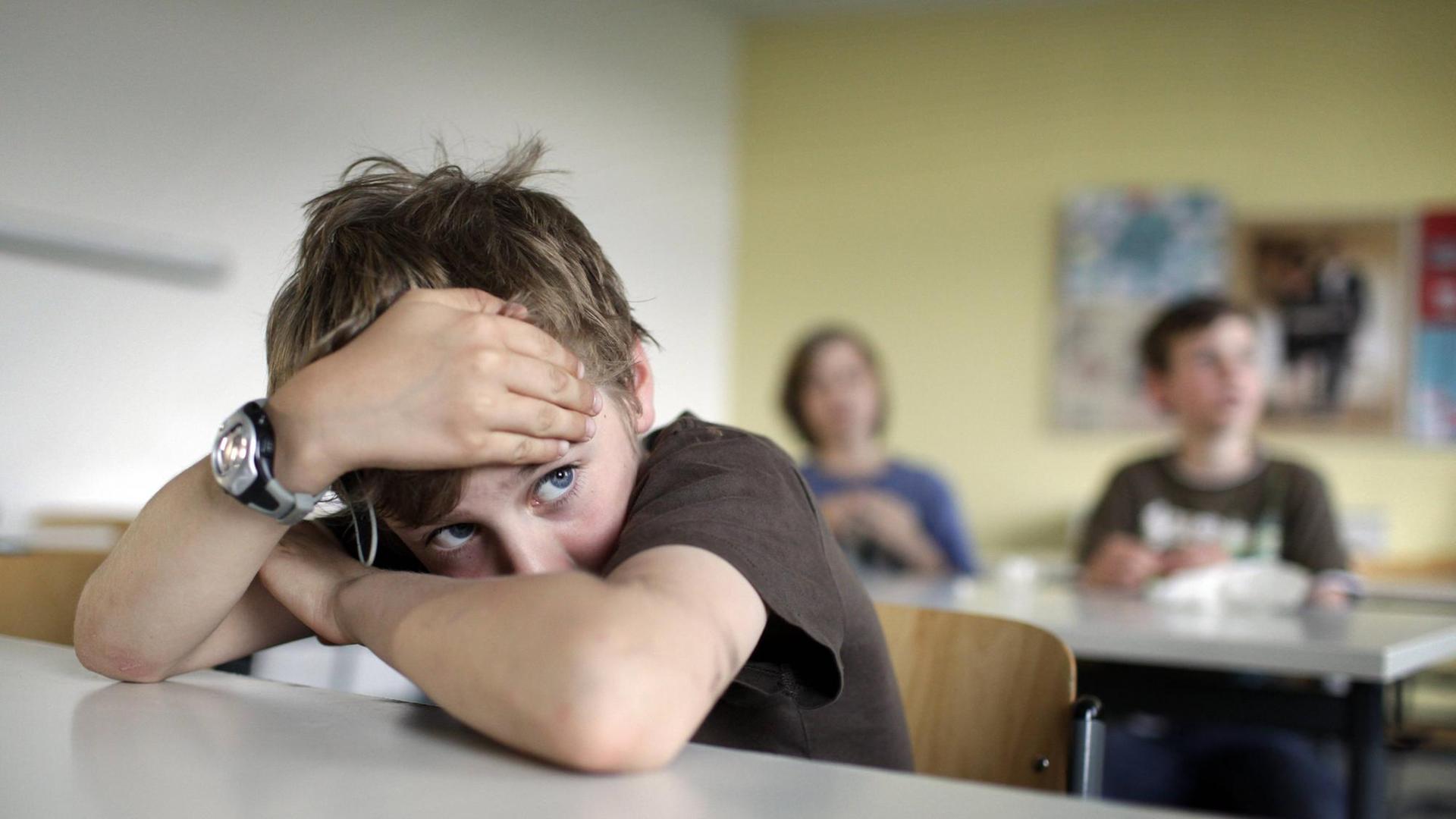 Ein Junge mit dunklem T-Shirt und zerzausten Haaren sitzt im Klassenzimmer und hat den Kopf auf die verschränkten Arme gestützt.