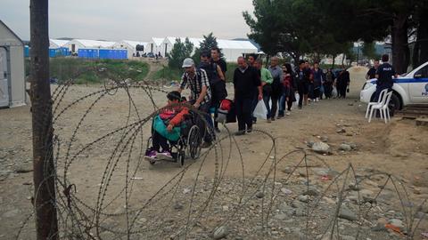 Syrische Flüchtlinge gehen hintereinander auf einen Stacheldrahtzaun zu. Ein Mann schiebt eine Frau im Rollstuhl.