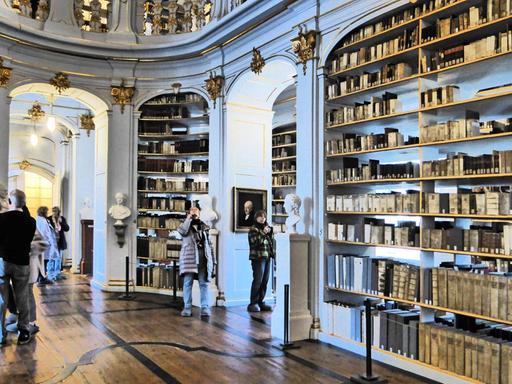 Blick in die Bibliothek mit Büchern auf Borden, stuckverzierten Wänden, Durchgängen und einer Empore