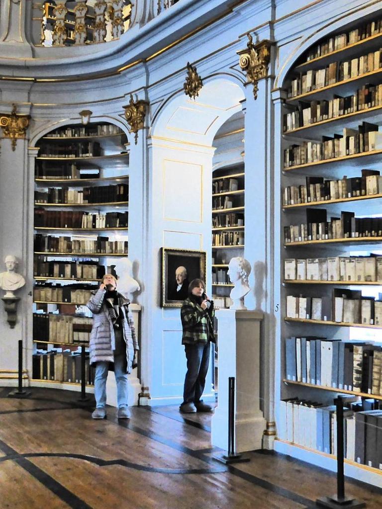 Blick in die Bibliothek mit Büchern auf Borden, stuckverzierten Wänden, Durchgängen und einer Empore