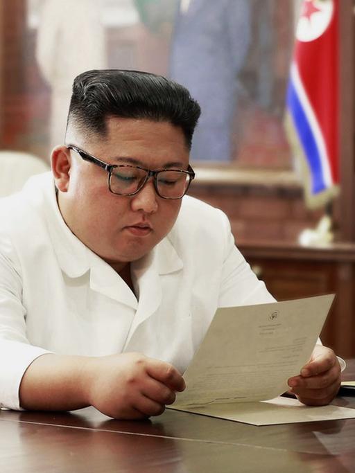 Kim sitzt im weißen Sakko in seinem Büro am Schreibtisch und liest den Brief.