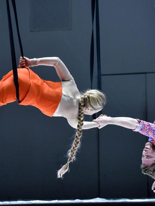 Links hängt eine Schauspielerin an einem Seil über der Bühne, sie hält rechts einen Schauspieler an der Hand, der kopfüber herunterhängt