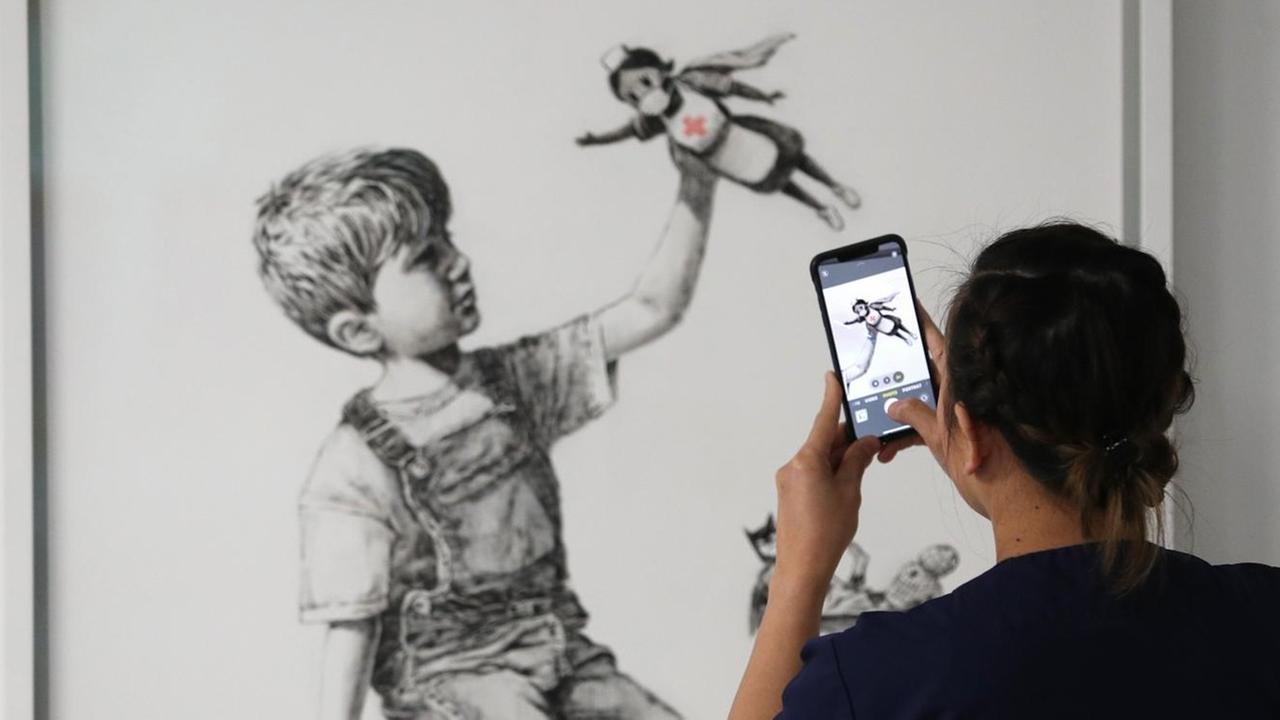 Eine Krankenschwester fotografiert mit ihrem Handy das neue Banksy-Kunstwerk: Es zeigt einen Jungen, der mit einer Krankenschwester als Superheldin spielt.