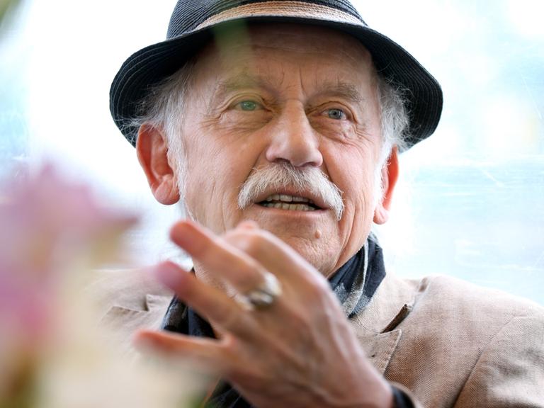 Tilo Prückner trägt einen Hut und gestikuliert.