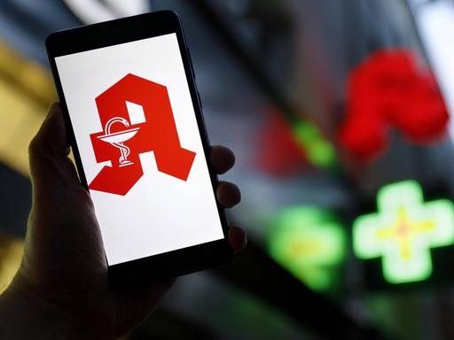 Das Apotheken-Logo erscheint auf einem Smartphone. Im Hintergrund ist eine Apotheke zu erkennen.