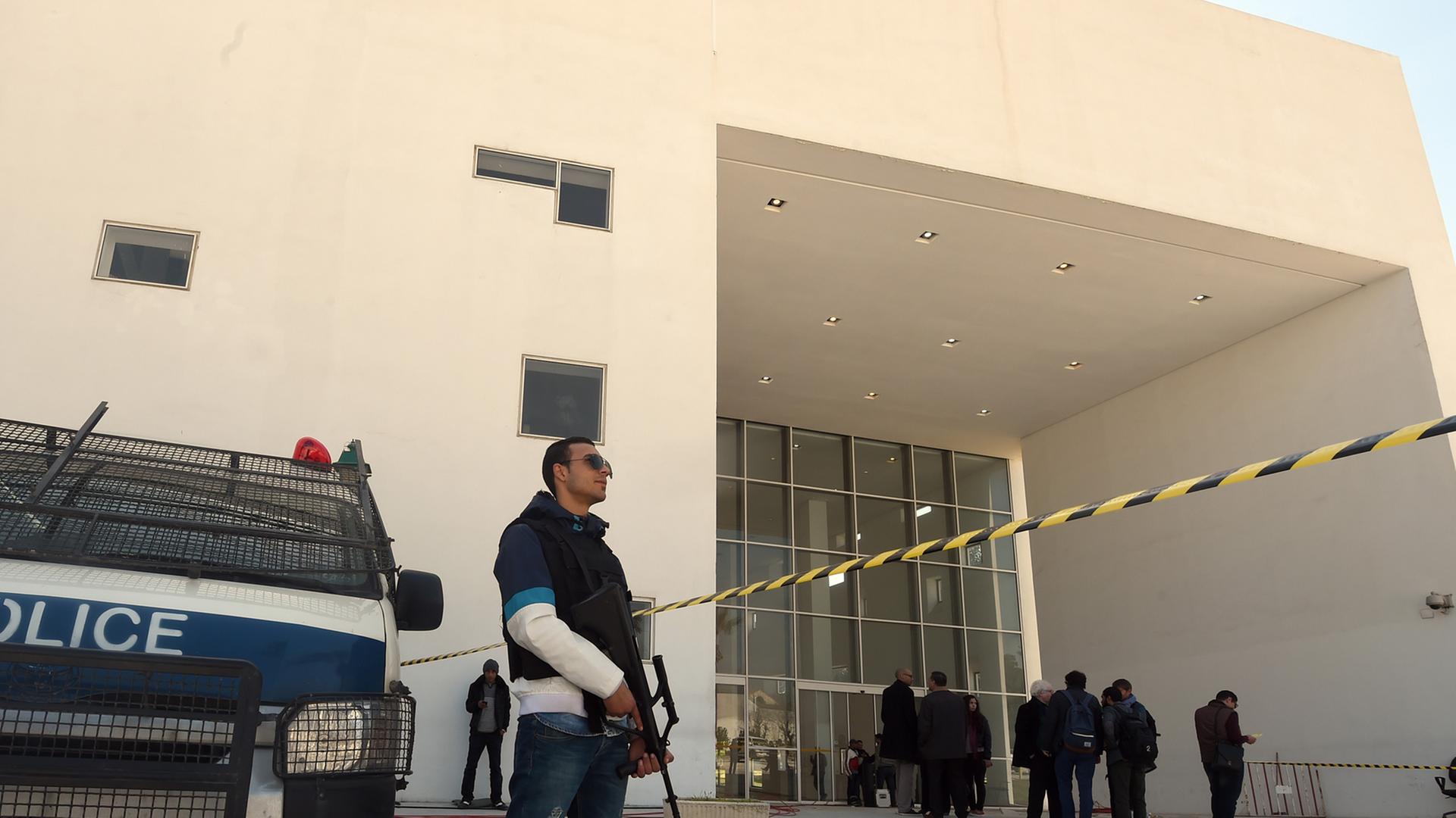 Ein Polizist steht neben einem gepanzerten Polizeifahrzeug vor dem Bardo-Museum in Tunis, dessen Eingangsbereich mit einem gelb-schwarzen Band abgesperrt ist. Im Hintergrund sieht man vor dem Eingang schwarzgekleidete Ermittler. 