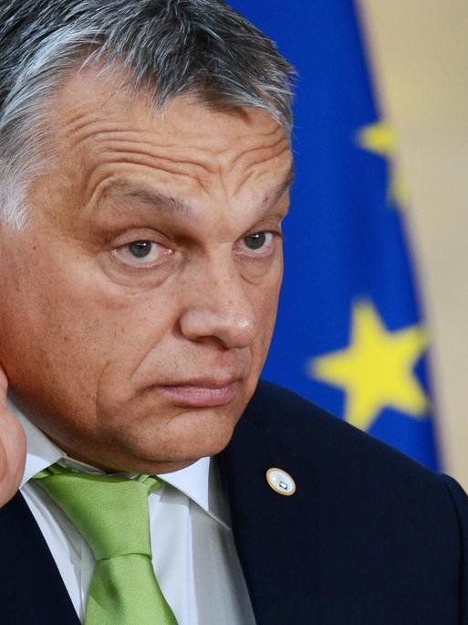 Viktor Orbán hält sich lauschend die rechte Hand hinter das Ohr, im Hintergrund eine EU-Fahne