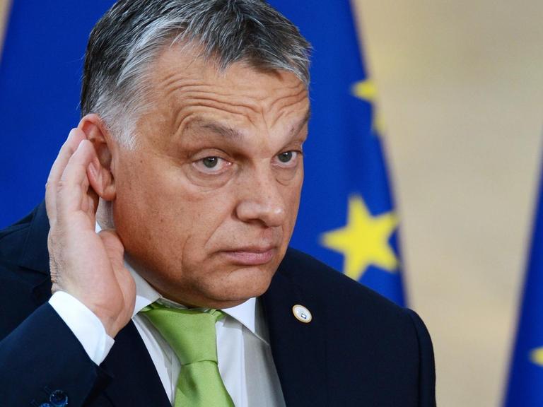 Viktor Orbán hält sich lauschend die rechte Hand hinter das Ohr, im Hintergrund eine EU-Fahne