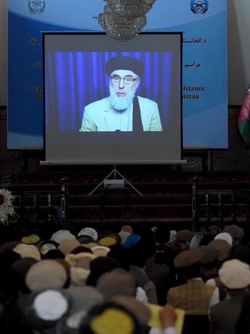 Auf einem Bildschirm in einem Saal ist der afghanische Kriegsherr Gulbuddin Hekmatyar zu sehen. Er unterschreibt gerade den Friedensvertrag mit der Regierung.