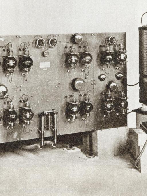 Schwarzweißaufnahme eines Radiosprechers der vor einer historischen Radioanlage sitzt.