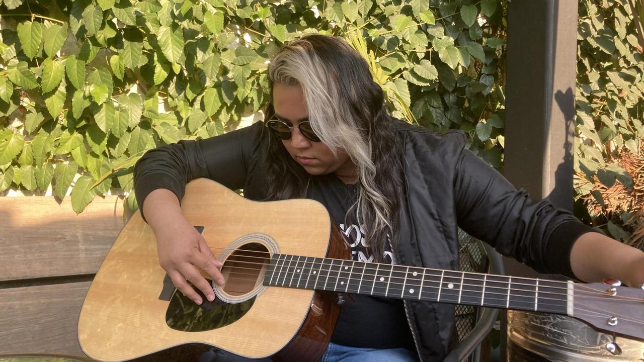 Eine junge Frau mit langen Haaren beugt sich auf einer Holzbank im Grünen über ihre Gitarre.
