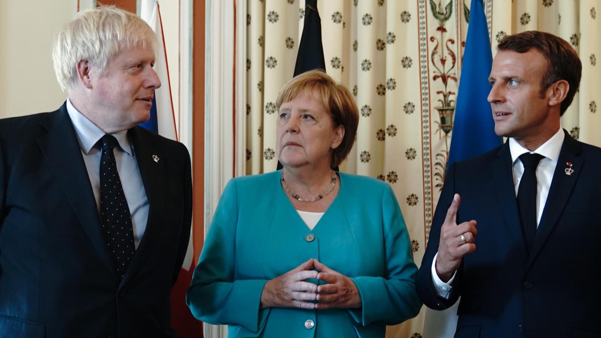 24.08.2019, Frankreich, Biarritz: Bundeskanzlerin Angela Merkel (CDU), steht zwischen Boris Johnson (l), Premierminister von Großbritannien, und Emmanuel Macron, Präsident von Frankreich