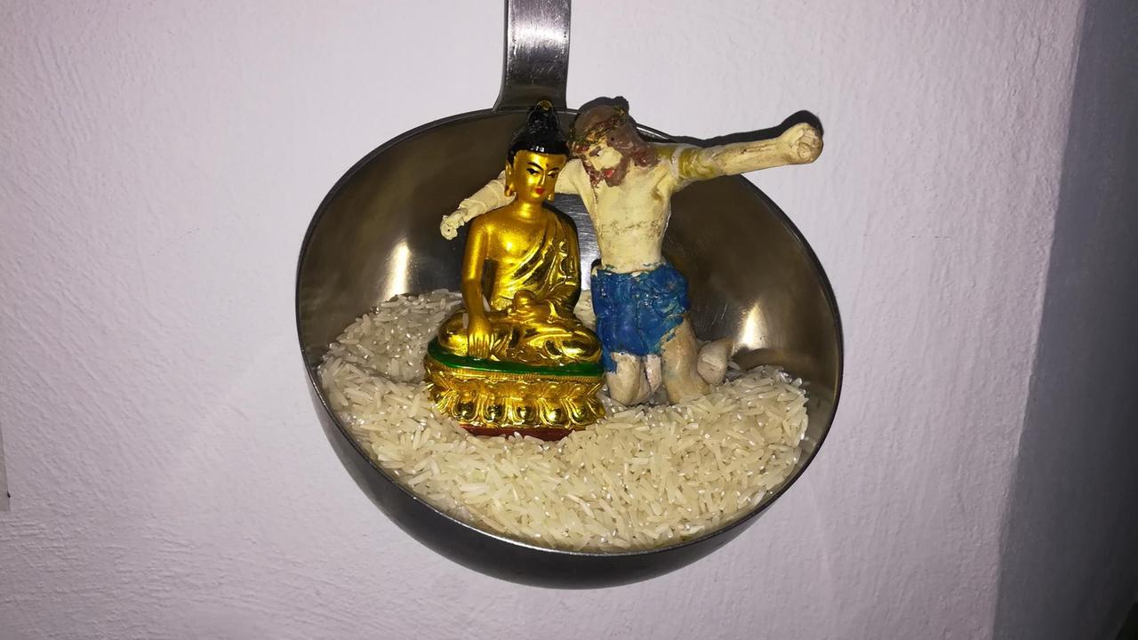 Das Bild zeigt das Kunstwerk "Brotherhood" von Hanne Draeger. Zu sehen sind eine Buddha-Statue und eine Jesus-Figur Arm in Arm in einer Suppenkelle voll Reis sitzend.