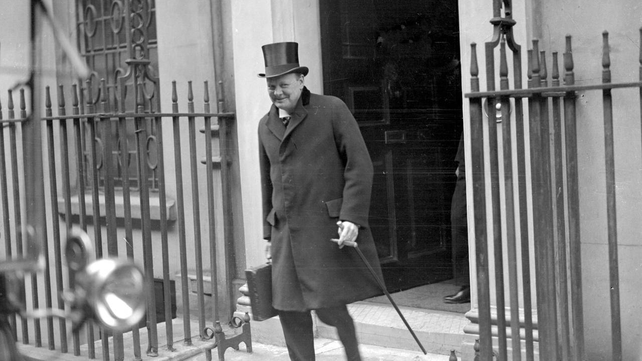Winston Churchill verlässt das Gebäude mit Hut und Gehstock. Er grinst verschmitzt in die Kamera.
