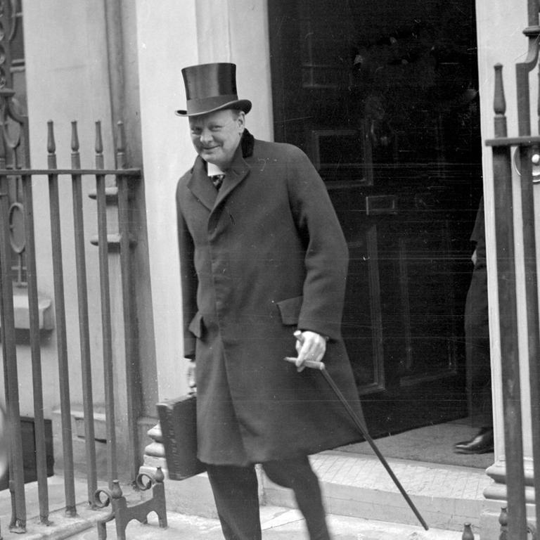 Winston Churchill verlässt das Gebäude mit Hut und Gehstock. Er grinst verschmitzt in die Kamera.