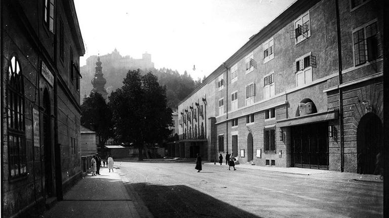 Auf einer alten Schwarzweißfotografie ist eine breite Straße zwischen massiven Häusern zu sehen, auf der nur wenige Menschen unterwegs sind.