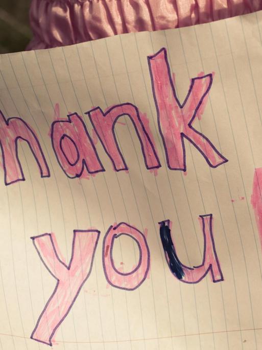 Ein kleines Mädchen in einem rosa Kleid hält ein selbstgemaltes Schild: 'thank you!'.