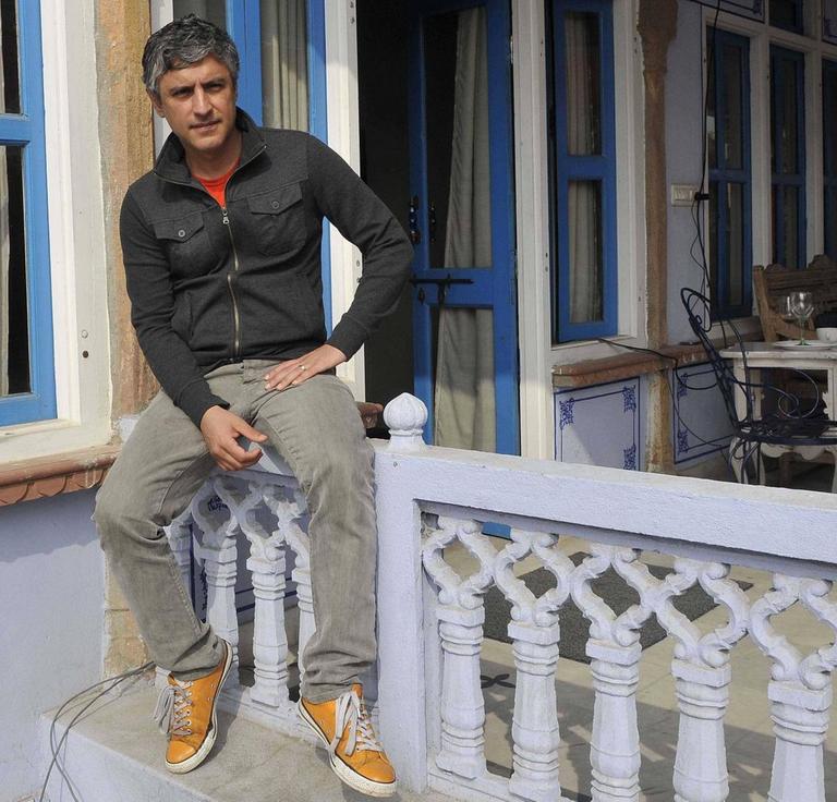 Reza Aslan sitzt in Indien auf einem Verandageländer vor einem Haus. 