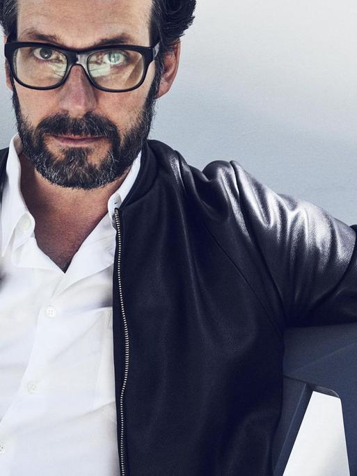 Porträt des Industrie-Designers Konstantin Cricic. Er trägt eine schwarze Lederjacke und sitzt in einem von ihm entworfenen Stuhl vor weißem Hintergrund.