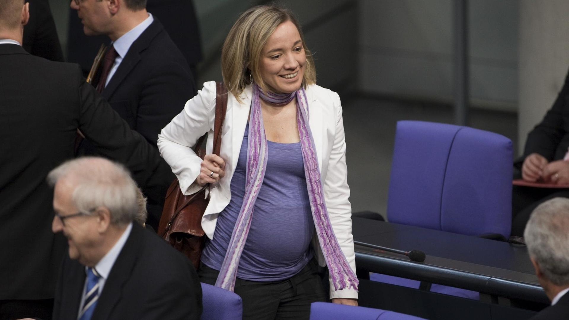 Eine hochwangere junge Frau geht durch die blauen Sitzreihen des Bundestags. Es befinden sich noch andere Politiker dort. Eine hochwangere junge Frau geht durch die blauen Sitzreihen des Bundestags. Es befinden sich noch andere Politiker dort.