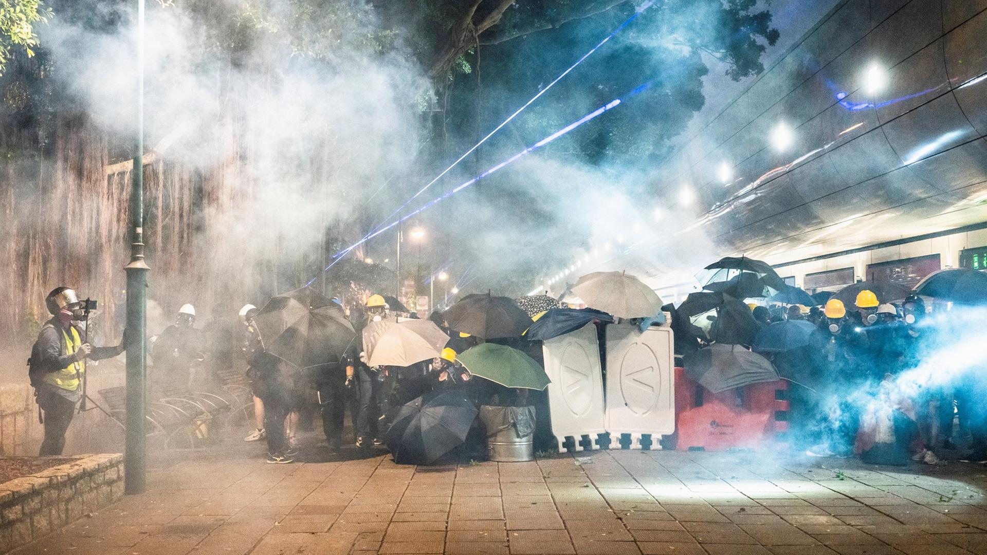 Demonstranten in Tsim Sha Tsui in Hongkong verstecken sich im Abendlicht hinter Regenschirmen und anderen Gegenständen vor dem sie umwabernden Tränengas.