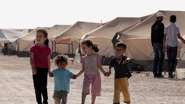 Vier Kinder, 2 größere Mädchen und 2 kleinere Jungs, gehen Hand in Hand auf einem Weg im Flüchtlingslager, im Hintergrund sind Zelte zu sehen