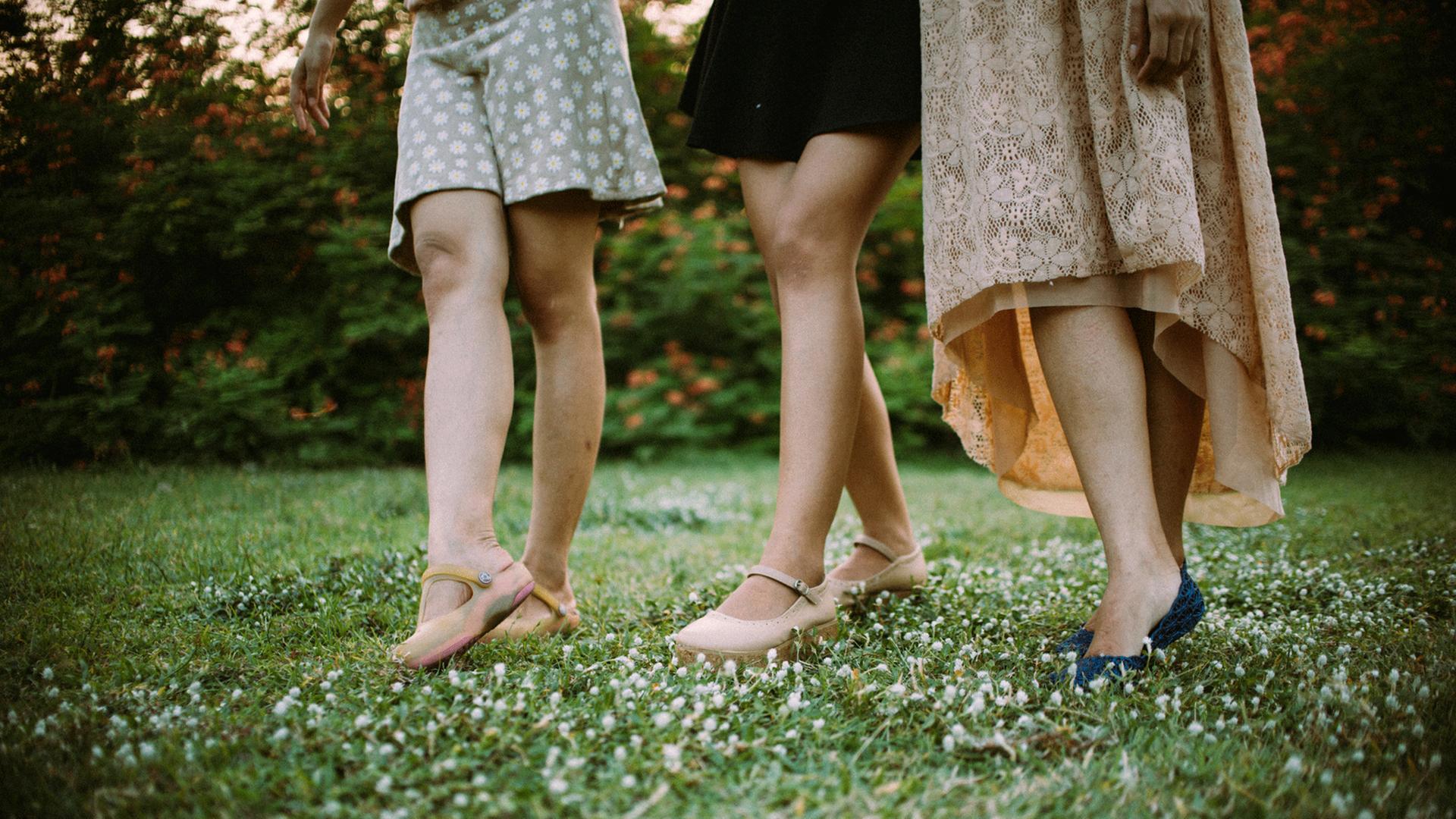 Die nackten Beine von drei Frauen in Röcken und Schuhen sind zu sehen. Sie spazieren über eine Wiese.