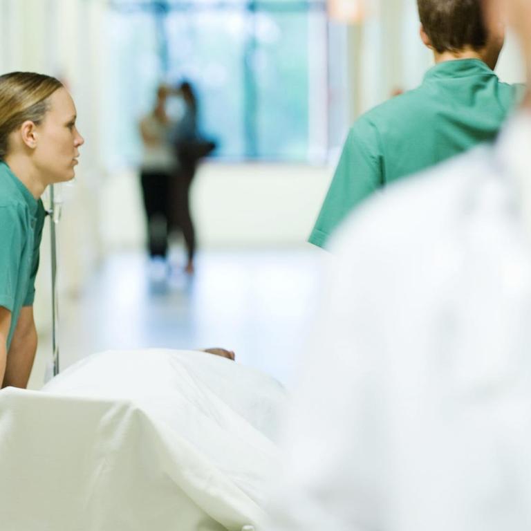 Ärztinnen schieben eine Krankenhausliege durch den Gang, ein Arzt läuft im Vordergrund durch das Bild