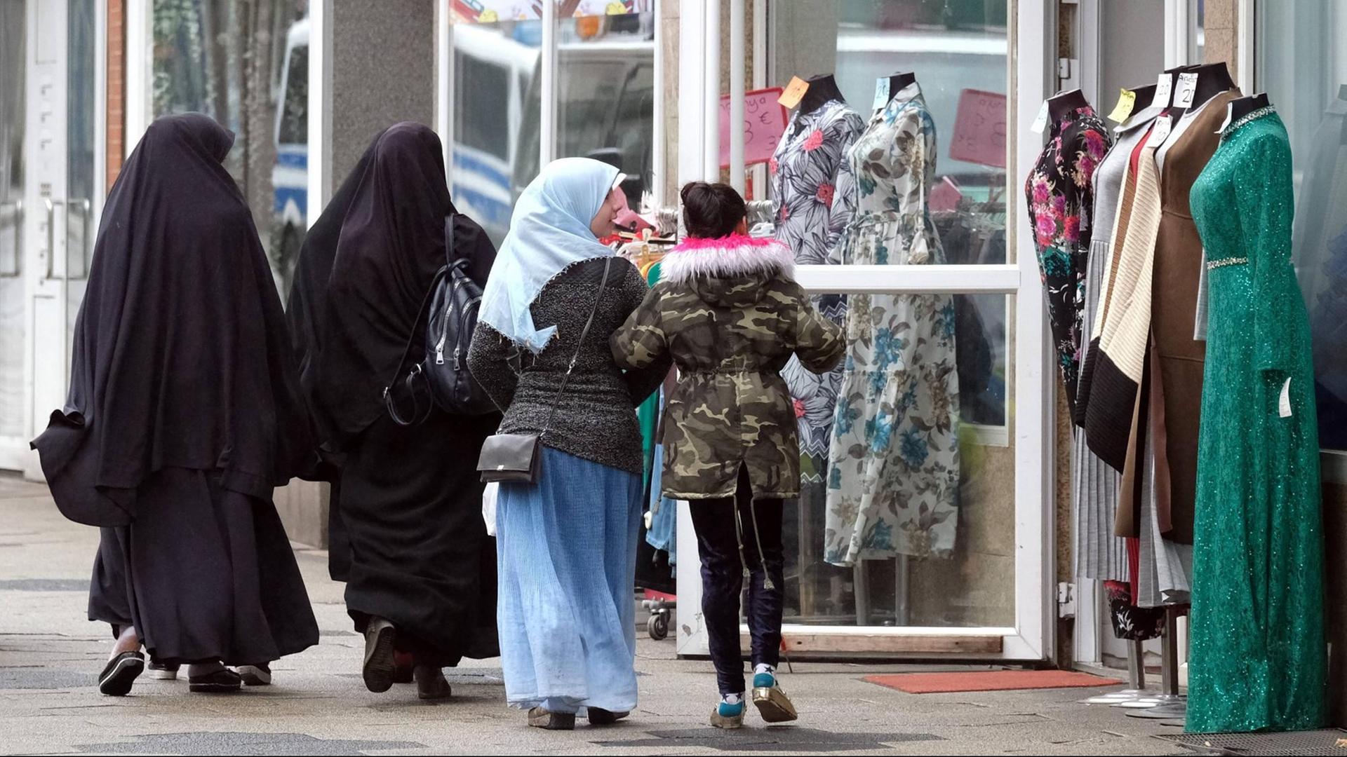 Zwei komplett in schwarz verhüllte ältere Frauen und zwei farbenfroher gekleidete jüngere Mädchen, davon eins mit mit buntem Kopftuch, passieren ein muslimisches Bekleidungsgeschaeft mit Schaufensterpuppen in Duisburg-Marxloh.