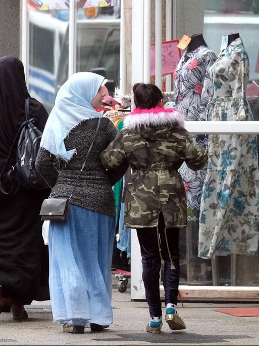 Zwei komplett in schwarz verhüllte ältere Frauen und zwei farbenfroher gekleidete jüngere Mädchen, davon eins mit mit buntem Kopftuch, passieren ein muslimisches Bekleidungsgeschaeft mit Schaufensterpuppen in Duisburg-Marxloh.