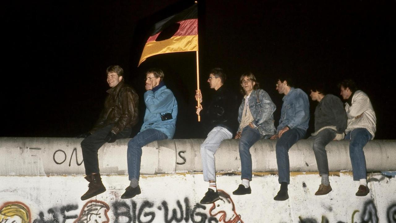 Ein Gruppe junger Männer sitzt mit einer zerschnittenen DDR-Fahne auf der Mauer am Brandenburger Tor in Berlin.