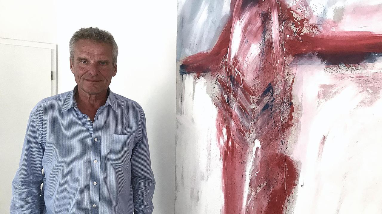 Der Theologe und Seelsorger Ronald Mundhenk neben dem Gemälde eines Psychiatrie-Patienten. Das Bild zeigt einen schemenhaften Körper, der an die Gestalt eines Gekreuzigten erinnert. 