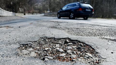 Das Bild zeigt ein Schlagloch auf einer Straße, im Hintergrund fährt ein Auto vorbei.