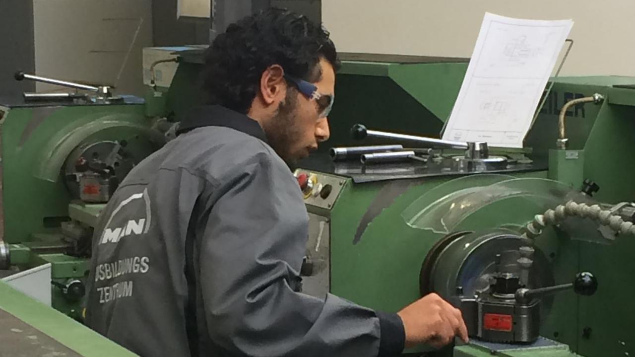 Der Auszubildende sitzt im grauen Overall und Schutzbrille einer grünen Maschine an einer Maschine.