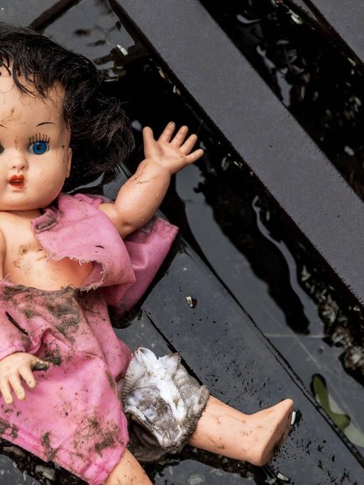 Eine Puppe liegt in dreckiger Bekleidung an einem Straßengully.