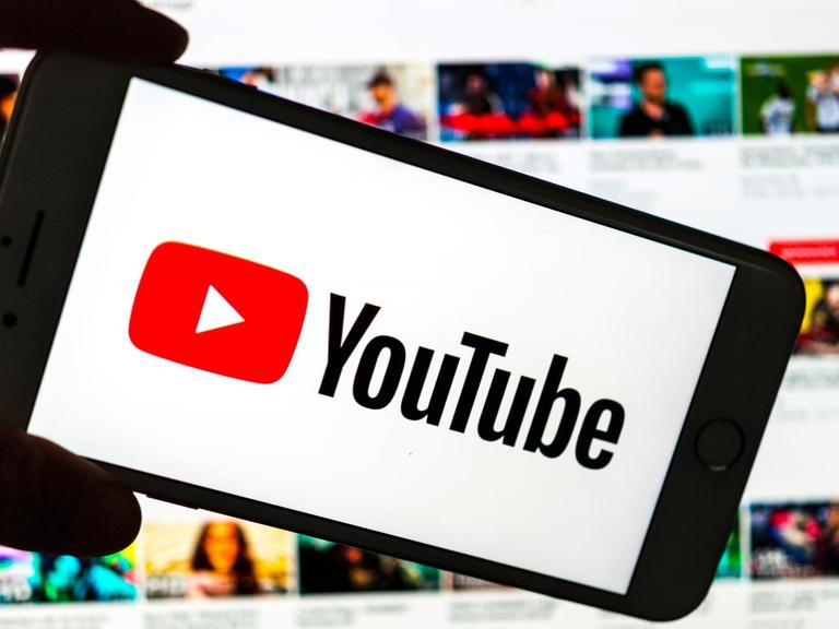 Das Logo des Video-Portals YouTube wird auf dem Display eines Smartphones angezeigt. Im Hintergrund ist auf einem Bildschirm die YouTube Homepage zu sehen.