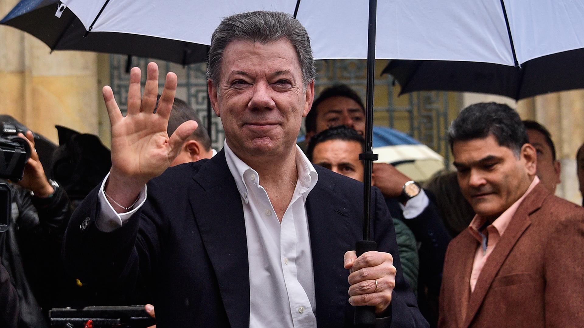 Der kolumbianische Präsident Juan Manuel Santos steht in einem dunklen Anzug und einem aufgespannten blauen Regenschirm mit mehreren Menschen zusammen.