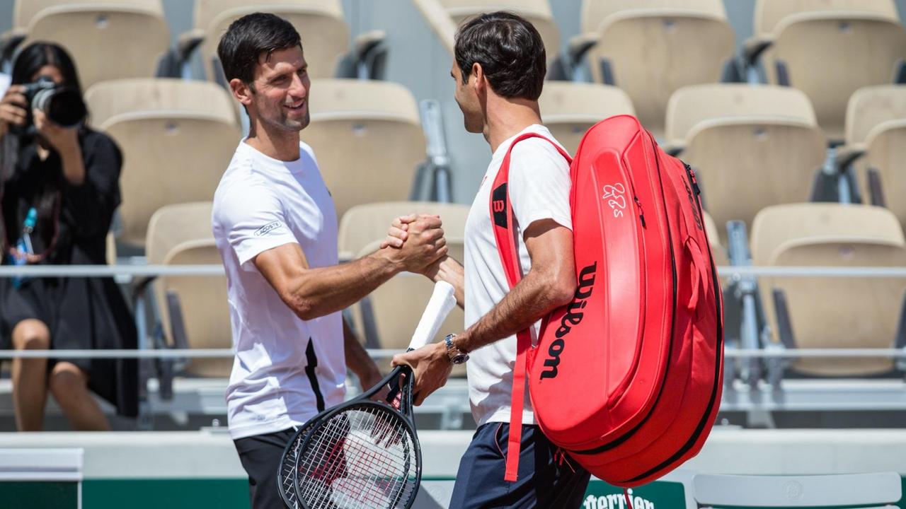 Man sieht Djokovic von vorne und Federer seitlich von hinten, beiden schütteln sich lächelnd die Hand. Im Hintergrund Zuschauersitze.